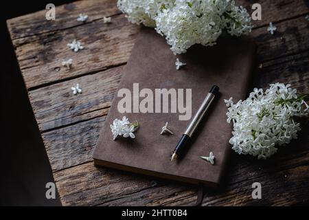 Spring romantic composition with white lilac flowers and old notebook for sketches on dark wooden background Stock Photo