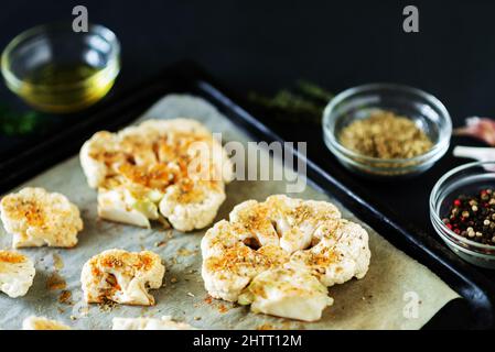 Cauliflower steak cooking. Raw cauliflower sprinkled with spices lies on a baking sheet. Olive oil, herbs, various spices nearby. Dark background. Stock Photo