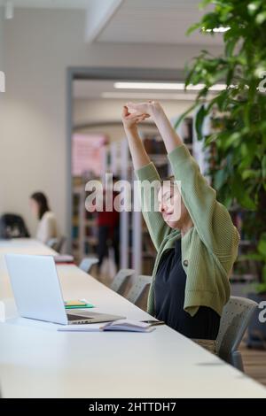 Tired woman adult learner stretching arms with closed eyes while studying in cozy library Stock Photo