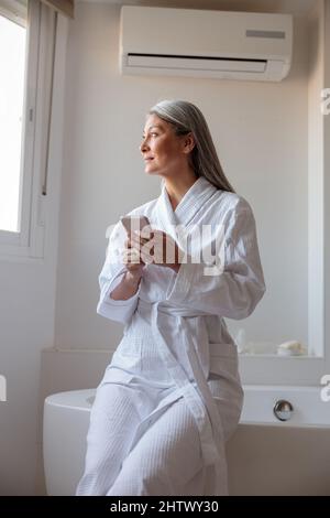 Pretty Asian woman in bathrobe holding smartphone in bathroom Stock Photo
