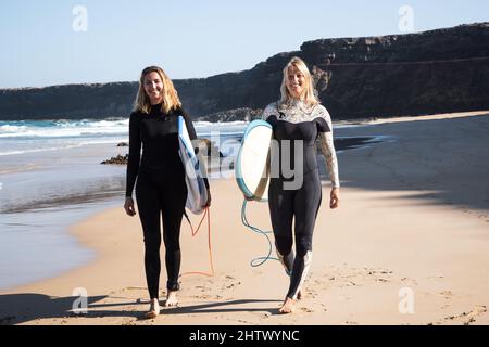 Surfer females walking on the beach shore. They are smiling and holding their surfboards. Stock Photo