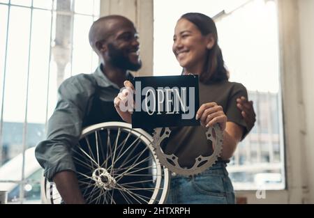 Your bike is in good hands here. Shot of a young couple standing together in their bicycle shop and holding an open sign. Stock Photo