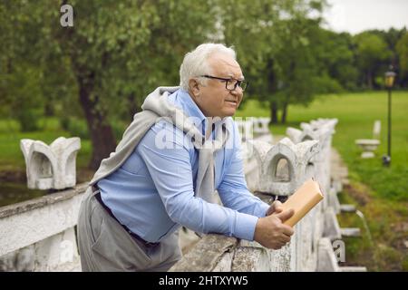 Pensive senior man holding book standing on bridge in park looking forward Stock Photo