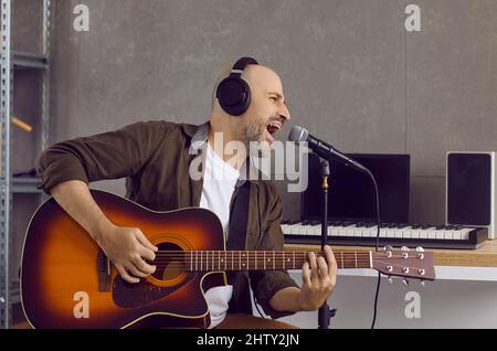 Portrait of man singing into microphone and playing guitar while recording music in studio. Stock Photo