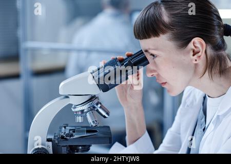 Female scientist in white coat sitting at table and analyzing the virus through the microscope while working at lab Stock Photo