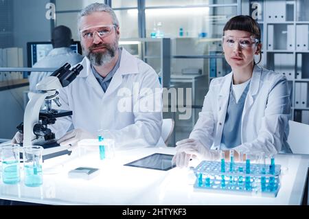 Portrait of two doctors in lab coats looking at camera while sitting at the table and working with samples in test tubes in team Stock Photo