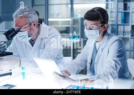 Female scientist in mask using tablet pc while working at the table with her colleague, they examining the dna of virus Stock Photo