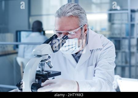 Mature scientist in mask looking through the microscope during scientific research in the laboratory, he working with samples of virus Stock Photo