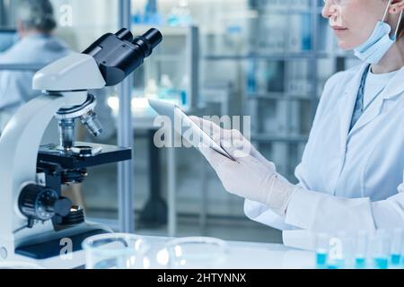 Close-up of woman in protective gloves and lab coat using digital tablet to examine the samples on the screen after researching them on microscope Stock Photo