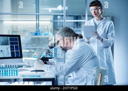 Mature scientist sitting at table and using microscope in medical exam with laboratory technician filling information on digital tablet Stock Photo