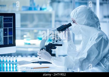 Female scientist in protective wear examining the sample of new virus through the microscope sitting at table in laboratory Stock Photo