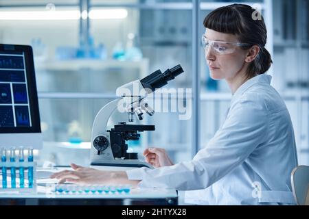 Female scientist sitting at table in laboratory and examining samples in test tubes with microscope Stock Photo