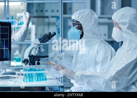 Two scientists in protective suits working with analysis in test tubes while sitting at the table with microscope in laboratory Stock Photo