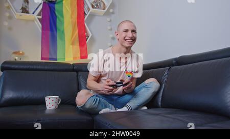 Happy queer non-binary person playing video games in their free time while sitting on a black sofa with rainbow pride flag behind their back. High quality photo Stock Photo
