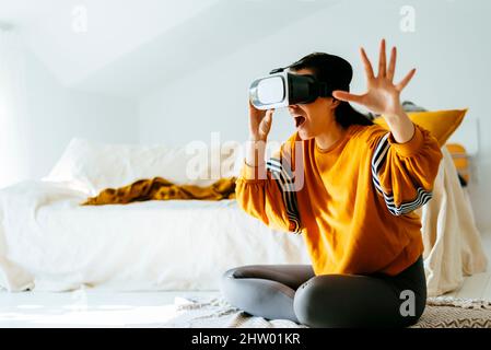 Excited casual clothing woman wearing virtual reality VR glasses headset in living room. Future trend using virtual reality goggles device for videoga Stock Photo