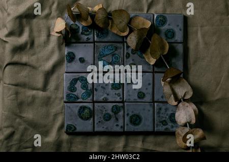 Dark black textured ceramic tiles blue spotted glaze on dark green cloth. Abstract background with eucalyptus branch. Top view, copy space Stock Photo