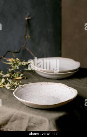 Set of empty craft ceramic white speckled bowls plates standing on dark green table cloth with dry branches Stock Photo