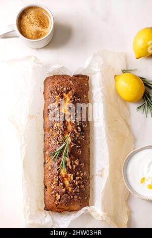 Whole Homemade lemon pound cake with nuts and rosemary on crumpled baking paper, serving with fresh lemon, white sauce and coffee mug on white marble Stock Photo
