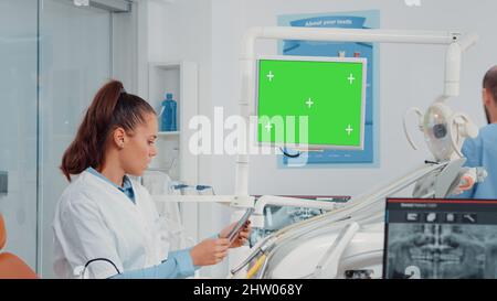 Dentist analyzing horizontal green screen on monitor and radiography of teeth for dental care. Woman using chroma key and mockup template for oral examination and teethcare in cabinet. Stock Photo