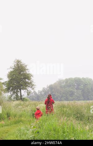 Mother with little child in red clothes walking through grassland. Light sky for copy space. Stock Photo