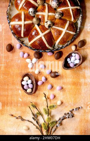Homemade Easter traditional hot cross buns in ceramic dish with blooming willow branches and chocolate eggs candies over orange stone background. Flat Stock Photo