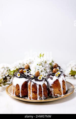 Homemade traditionla Easter kulich cake with chocolate nests and eggs on ceramic plate and blossoming cherry tree over white texture background. Tradi Stock Photo