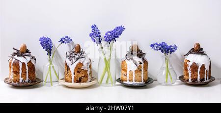 Homemade traditionla small Easter kulich cakes with chocolate nests and eggs on plates in row decorated with muscari flowers over white marble table. Stock Photo