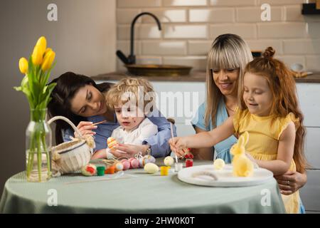 Two mothers with their children coloring Easter eggs while sitting in the kitchen Stock Photo