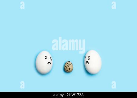 Two eggs with angry face looking at a quail egg on a blue background Stock Photo