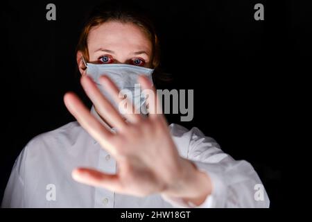 A scented woman with red painful eyes makes a warning stop gesture with her hand. Portrait of a woman on a black background during the coronavirus epi Stock Photo