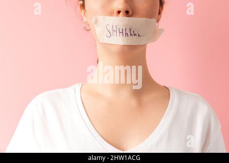 Close up of caucasian woman's face with her mouth taped shut. Tape has the inscription Shhh. Pink background. The concept of women's secrets and probl Stock Photo