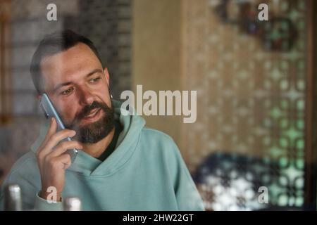 Young businessman talking on his phone indoor through glass. Bearded man in casual clothes using his smartphone. Stock Photo