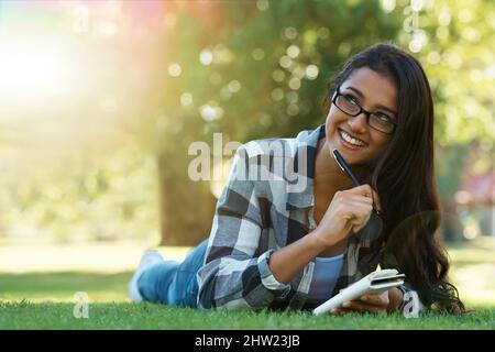 Jotting down her thoughts. A young woman lying on the grass writing in a notebook. Stock Photo