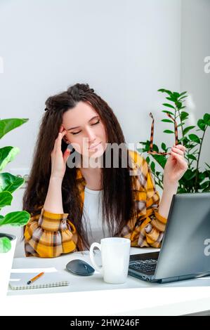 Beautiful woman holds her temples because of a headache or eye pain from working at a laptop. Woman working at home, undergoing training, watching web Stock Photo