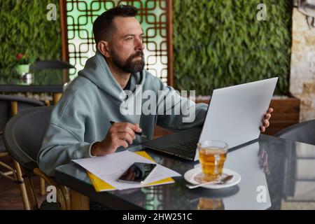 Thoughtful bearded man in casual clothes in cafe using laptop. Online business Stock Photo