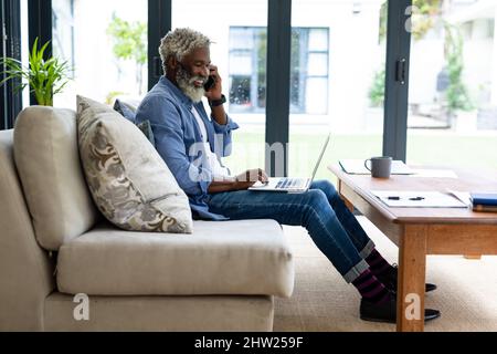 Smiling african american senior businessman talking mobile phone using laptop on sofa in living room Stock Photo