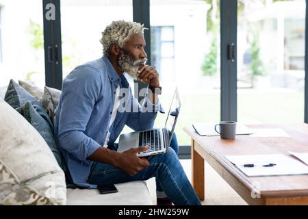 Thoughtful african american male freelancer with laptop sitting on sofa in living room at home Stock Photo