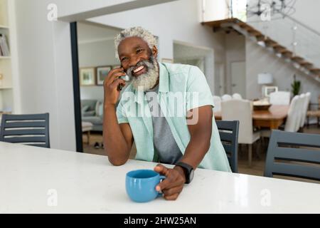 Happy senior african american man talking mobile phone while holding coffee mug in kitchen at home Stock Photo