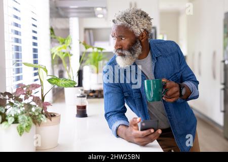 African american man holding coffee mug and smartphone looking away while leaning on kitchen counter Stock Photo