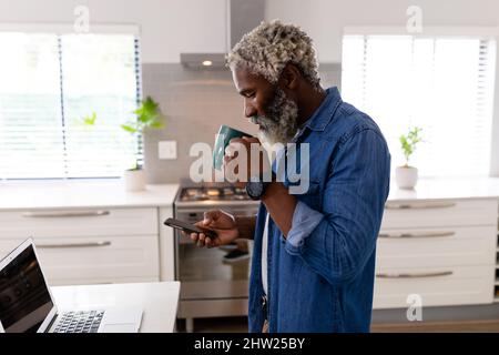 African american man drinking coffee from mug while using mobile phone in kitchen at home Stock Photo