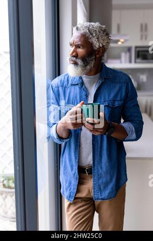 Bearded african american senior man holding coffee mug leaning by window in kitchen at home Stock Photo