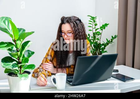 Smiling woman in glasses writing in a notebook on a workplace. Woman working at home, takes notes at a meeting, undergoing training, watching webinar Stock Photo