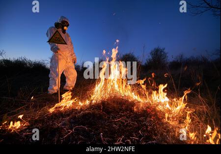 Fireman ecologist extinguishing fire in field at night. Man in suit and gas mask near burning grass, holding yellow triangle with skull and crossbones warning sign. Natural disaster concept. Stock Photo