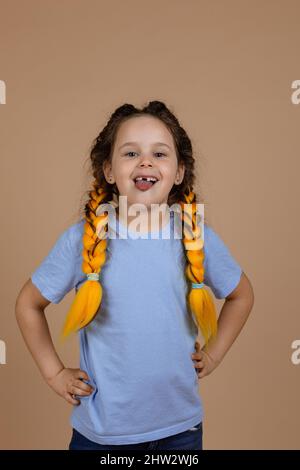 Mischievous little shining girl with slight smile showing tongue looking at camera misbehaving having kanekalon braids on beige background wearing Stock Photo