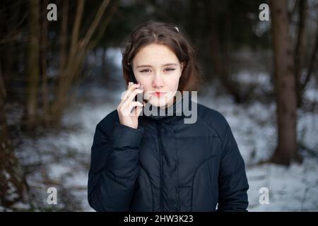 Girl is talking on phone in woods. Schoolgirl is alone in park in winter. Woman holds smartphone near her head. Walk in cold weather. Tense conversati Stock Photo