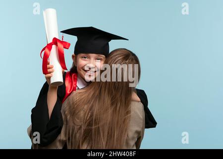 Little girl graduate celebrating graduation. Child wearing graduation cap and ceremony robe Holding Certificate. Mom hugs and congratulations daughter on graduation. Successfully complete course study Stock Photo