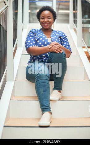 Making my way to the top one step at a time. Portrait of a confident young businesswoman sitting on the steps of a modern office. Stock Photo