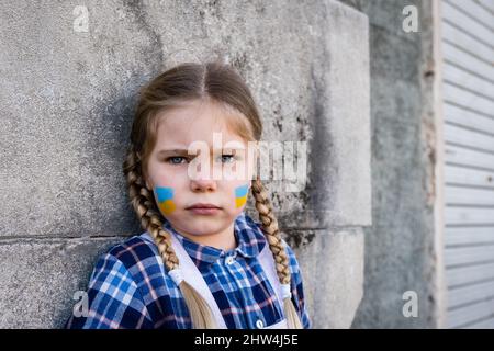 Portrait of a frightened child girl with painted flag on the cheek in yellow-blue colors of the Ukrainian flag. Peace and protection children concept Stock Photo