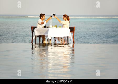Happy Anniversary. A happy couple clinking their wine glasses while sitting outside surrounded by water. Stock Photo