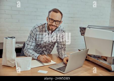 Cheerful man using laptop and taking notes in cafe Stock Photo